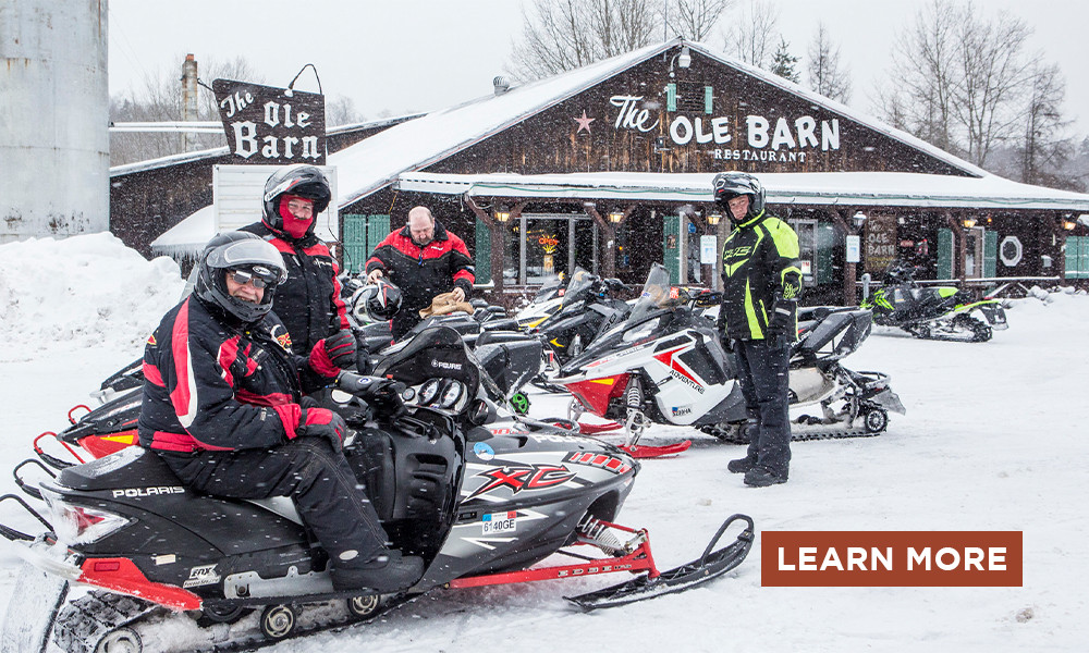 A group of snowmobilers pose on their sleds in front of a rustic restaurant on a snowy day.