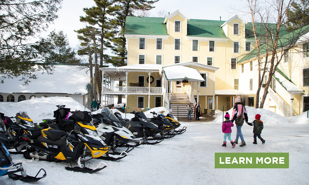 A woman and two children walk past a row of snowmobiles in front of a large Adirondack inn.