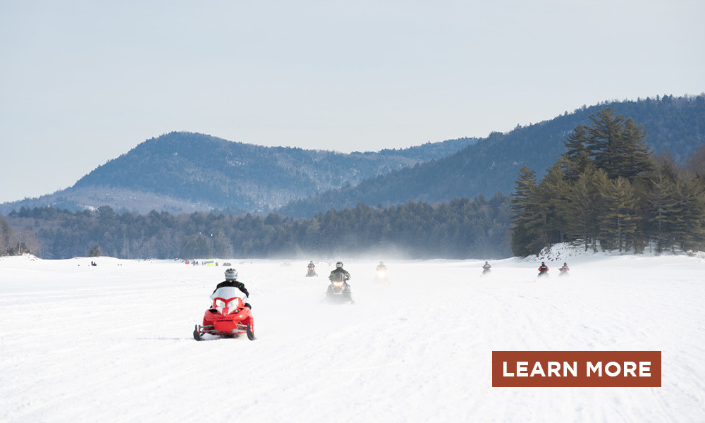 Snowmobilers zoom across a frozen lake with low mountains beyond.