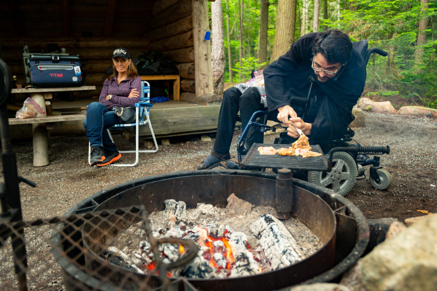 A man in a wheelchair cooks with an accessible grill in front of a lean-to.